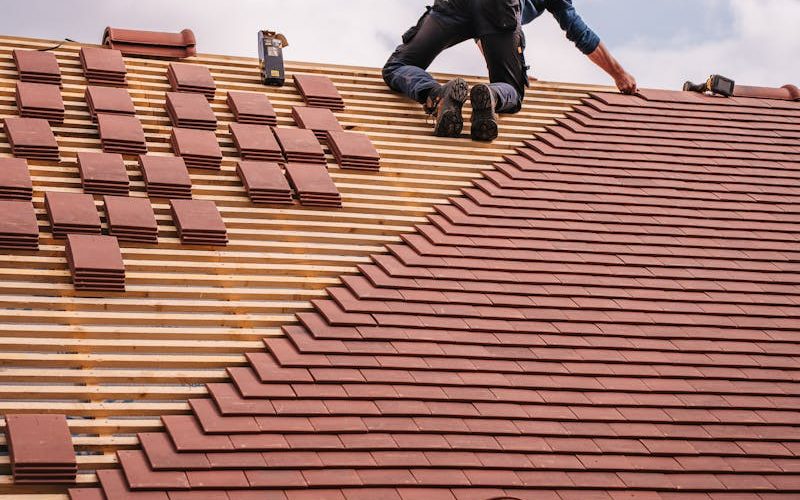 A roofer carefully installs shingles on a new residential roof under a cloudy sky.